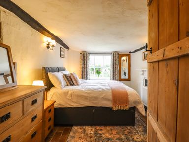 A bedroom with a bed, wooden dresser, window with curtains, and a mirror on the wall at The Abbey Cottage in Windsor Green