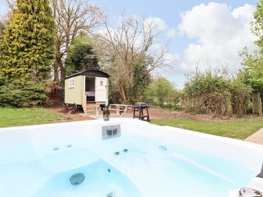 An outdoor scene with a hot tub in the foreground a small green wooden hut with a white door and steps picnic table and barbecue grill in the background at The Abbey Cottage in Windsor Green
