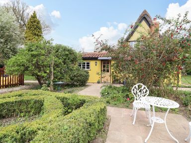 A garden with trimmed hedges a white metal table and chair and a yellow house with shrubs at The Abbey Cottage in Windsor Green