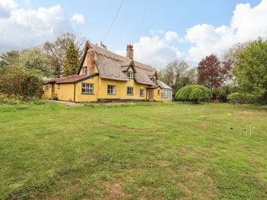 A yellow thatched roof house with a large lawn and trees at The Abbey Cottage in Windsor Green