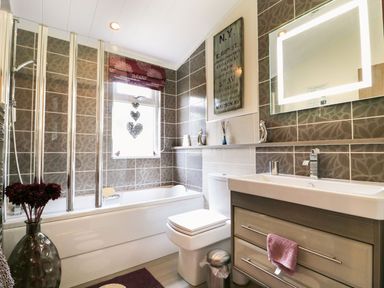 A bathroom with a bathtub with shower screen a toilet a sink with cabinet a lit mirror and decorative wall art at Hafan Lodge 85 Wisemans Bridge near Saundersfoot