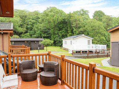 A wooden deck with two round wicker chairs and a small table overlooking a green lawn with cabins at Hafan Lodge 85 Wisemans Bridge near Saundersfoot