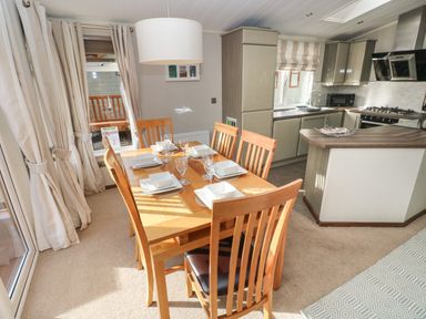 A dining area with a wooden table and six chairs set with white dishes next to a kitchen with gray cabinets and a window at Hafan Lodge 85 Wisemans Bridge near Saundersfoot