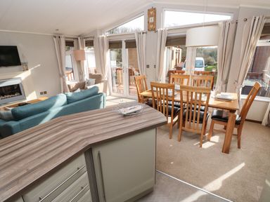 An open plan living and dining area with a kitchen counter a blue sofa a wooden dining table with six chairs and large windows at Hafan Lodge 85 Wisemans Bridge near Saundersfoot