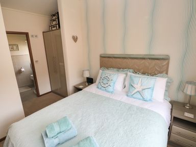 A bedroom with a double bed with light blue bedding and decorative pillows a bedside table with a lamp and an open door revealing a bathroom at Hafan Lodge 85 Wisemans Bridge near Saundersfoot