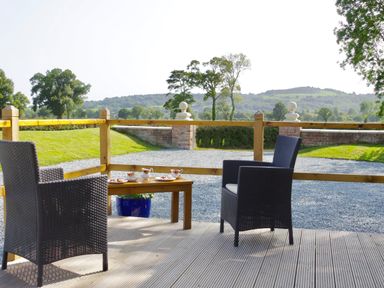 A wooden deck with two dark wicker chairs and a small table set with tea cups and plates with a gravel driveway and trees in the background at Haimwood Byetake in Llandrinio