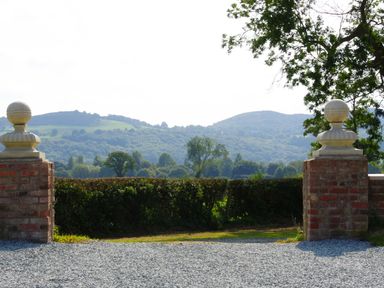 A gravel driveway entrance with brick pillars and stone finials overlooking green hills and trees at Haimwood Byetake in Llandrinio