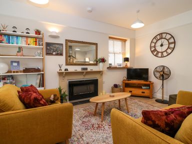 A living room with yellow sofas a coffee table a television a wall clock and bookshelves at Hardings cottage in Minehead