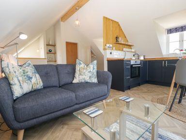 A living room area with a blue sofa and patterned cushions next to a glass coffee table and an open kitchen with dark cabinets and wooden accents at Little Knaplock in Fiddington near Combwich Somerset