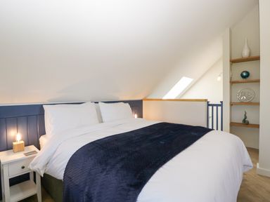 A bedroom with a double bed white bedding and navy blue throw next to a nightstand with a lamp and wall shelves with decorative items at Little Knaplock in Fiddington near Combwich Somerset