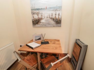 A small dining area with a wooden table and two chairs a folding chair leaning against the wall and a beach painting on the wall at Coras House in Amble