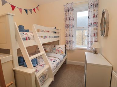 A small bedroom with a bunk bed and a window with curtains a white cabinet with a lamp and a decorative wall piece at Coras House in Amble
