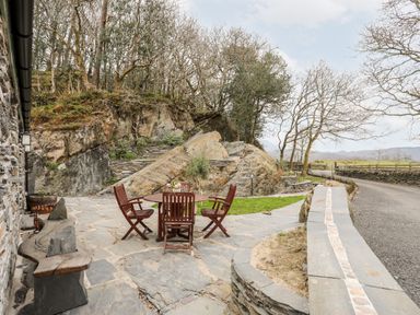 An outdoor stone patio with a round wooden table and four chairs near a stone building and rocky hillside at Tyn Llwyn Cornel Eco Barn in Garreg