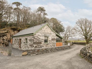 A stone-built barn with a gravel driveway and leafless trees at Tyn Llwyn Cornel Eco Barn in Garreg