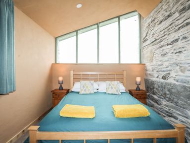 A bedroom with a wooden bed frame blue bedding and yellow towels on the bed two bedside tables with lamps and a stone wall at Tyn Llwyn Cornel Eco Barn in Garreg