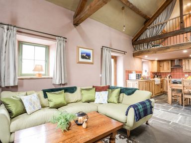 A living room with a green sectional sofa and wooden coffee table next to a kitchen with wooden cabinets at Tyn Llwyn Cornel Eco Barn in Garreg