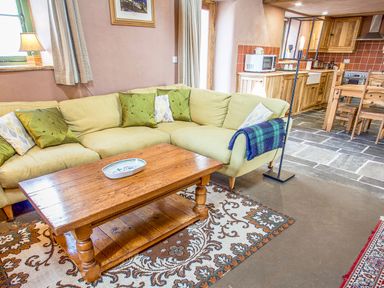 A living room with a yellow corner sofa with green and patterned cushions a wooden coffee table on a patterned rug next to a kitchen with wooden cabinets at Tyn Llwyn Cornel Eco Barn in Garreg