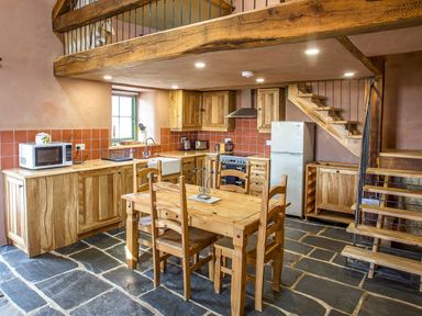 A kitchen with wooden cabinets a dining table four chairs a stove refrigerator microwave and staircase at Tyn Llwyn Cornel Eco Barn in Garreg
