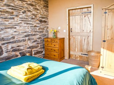 A bedroom with a blue bedspread and yellow towels on the bed a wooden dresser with flowers a wooden door a wicker basket and a wooden wardrobe at Tyn Llwyn Cornel Eco Barn in Garreg