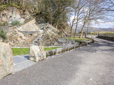 A gravel driveway with large rocks and stone steps leading up to a rocky hillside with trees and grassy areas at Tyn Llwyn Cornel Eco Barn in Garreg
