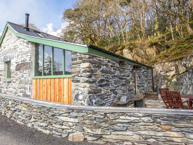 An exterior view of a stone house with green framed windows and wooden panels next to a stone wall and outdoor wooden furniture at Tyn Llwyn Cornel Eco Barn in Garreg