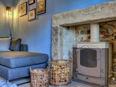 A living area with a gray sofa next to a stone fireplace with a wood stove and wicker baskets filled with firewood at Crab Cottage in Sandsend
