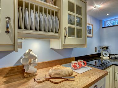 A kitchen countertop with a loaf of bread and knife two mugs with lobsters a plate rack with plates white hanging cups a kitchen scale and an electric stove at Crab Cottage in Sandsend