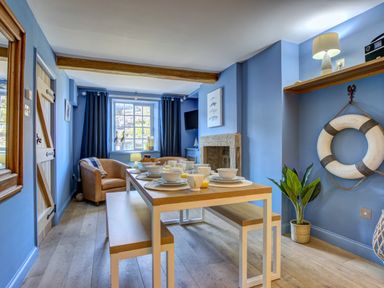 A dining area with a wooden table set with bowls and cups two benches a brown armchair a window with dark curtains a fireplace a wall shelf with a lamp and a plant in a pot at Crab Cottage in Sandsend