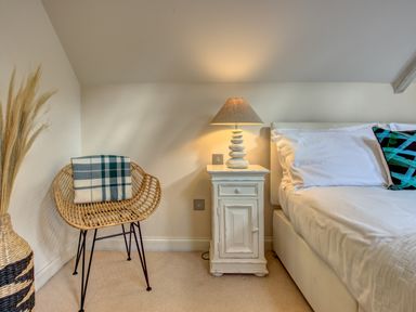 A bedroom corner with a wicker chair holding a plaid blanket a bedside cabinet with a lamp and part of a bed with white linen at Crab Cottage in Sandsend