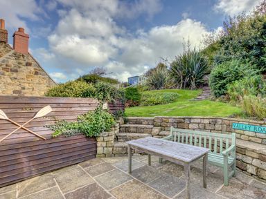 An outdoor patio with a wooden table and bench next to stone walls and stairs leading to a grassy garden at Crab Cottage in Sandsend