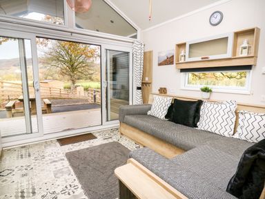 A living room with a gray sofa and patterned pillows near sliding glass doors with a view of a tree at Oak Tree View in Nantmel near Rhayader