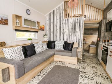 A living room with a gray and wood sofa with black and white pillows a patterned rug and a small kitchen at Oak Tree View in Nantmel near Rhayader