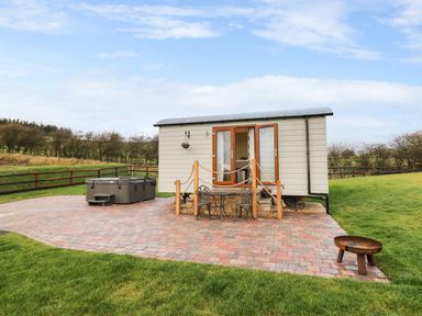 A small cabin with a patio table and chairs and a hot tub on a paved area with a fire pit on grass at Highland View in Rhayader