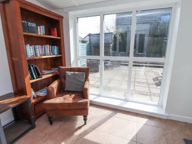 A room with a brown armchair a wooden bookshelf and a small wooden table by large windows at The Old School in Lowick