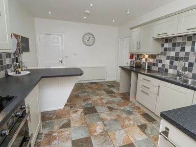A kitchen with tiled floor white cabinets black countertops a wall clock and a bottle with glasses on the counter at The Old School in Lowick