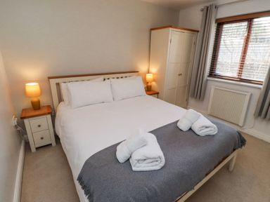 A bedroom with a bed with white sheets and gray blanket with two folded towels on top two bedside tables with lamps a white wardrobe and a window with blinds and curtains at The Old School in Lowick