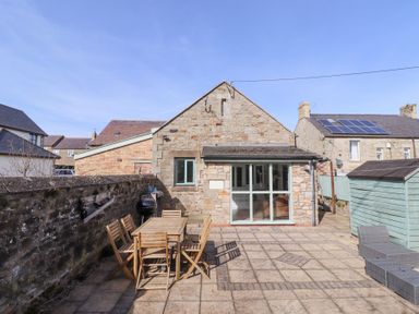 A stone house with a patio featuring a wooden table and chairs a barbecue and a garden shed at The Old School in Lowick