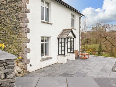 An outdoor patio area with stone tiles beside a white house with stone corner accents and wooden bench with a table at Estcote in Roger Ground near Hawkshead