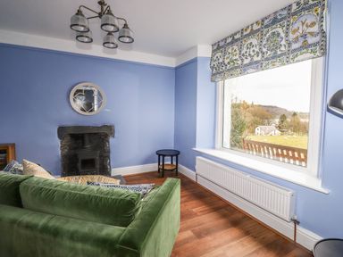 A sitting room with a green sofa a small round table a fireplace and a large window with a floral curtain at Estcote Roger Ground near Hawkshead