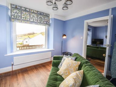 A room with a green couch with patterned pillows a floor lamp a window with a floral valance and wooden floor at Estcote in Roger Ground near Hawkshead