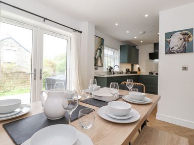 A dining table set with white dishes and glassware near glass doors and a kitchen with dark cabinets at Rydal Rock in Ambleside