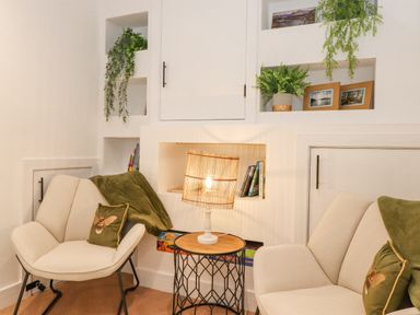 A seating area with two beige chairs a wooden side table with a lamp built in shelves with plants photos and books at Rydal Rock in Ambleside