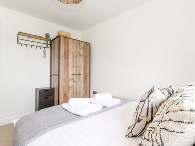 A bedroom with a bed folded towels a wooden wardrobe and a small drawer cabinet with a wall shelf