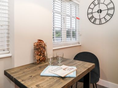 A wooden table with two glasses and books next to a window with blinds and a black chair in a room with a wall clock at Fenty's in Bowness-On-Windermere