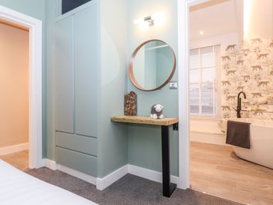A corner of a bedroom with a built-in wardrobe a small wooden table with a round mirror above it and an adjacent bathroom with a bathtub and patterned wallpaper at Fenty's in Bowness-On-Windermere