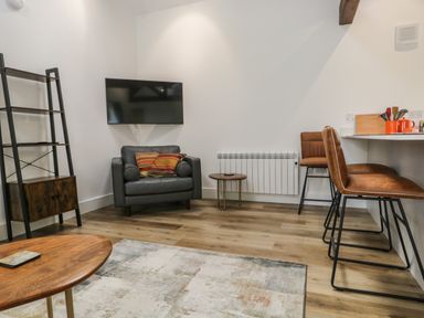 A living room with a grey armchair and cushions a wall mounted television a wooden shelf a small round table and two brown bar stools at Bank Hurst Coach House in Ulverston