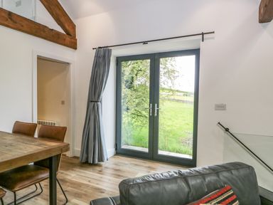 A room with wooden dining table and chairs near glass doors with curtain and a black leather sofa at Bank Hurst Coach House in Ulverston