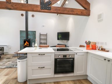 A kitchen with a white counter and built-in oven next to a living area with a wood burning stove and a wall-mounted television at Bank Hurst Coach House in Ulverston