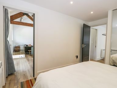 A bedroom with a white bed facing an open door to a living area with exposed ceiling beams at Bank Hurst Coach House in Ulverston