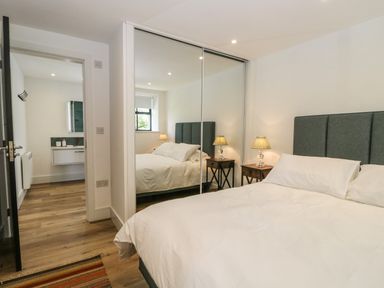 A bedroom with a bed a bedside table with a lamp a mirrored wardrobe and a doorway leading to a bathroom at Bank Hurst Coach House Ulverston